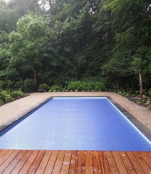 Afgewerkt zwembad met blauwe lamellenafdekking en houten vlonder, met bomen en struiken erachter.