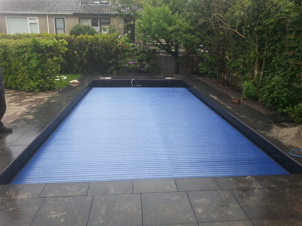 Zwembad met gesloten blauwe lamellenafdekking en donkere randafwerking, zand en haag rondom.