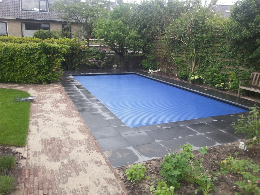 Zwembad met blauwe lamellenafdekking, donkere tegelrand en klinkerpad in een besloten tuin in Soest.