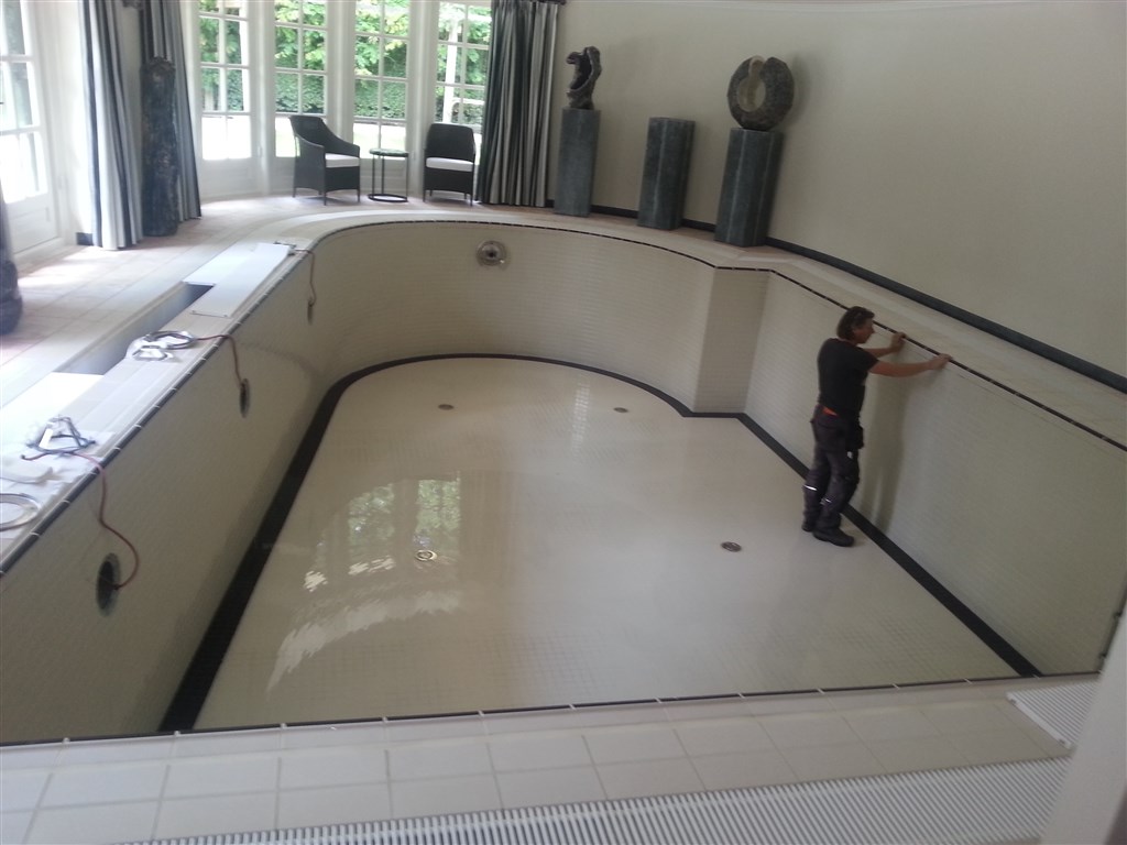 Leeg binnenzwembad met lichte wanden en zwarte band, terwijl een man aan de wand werkt.