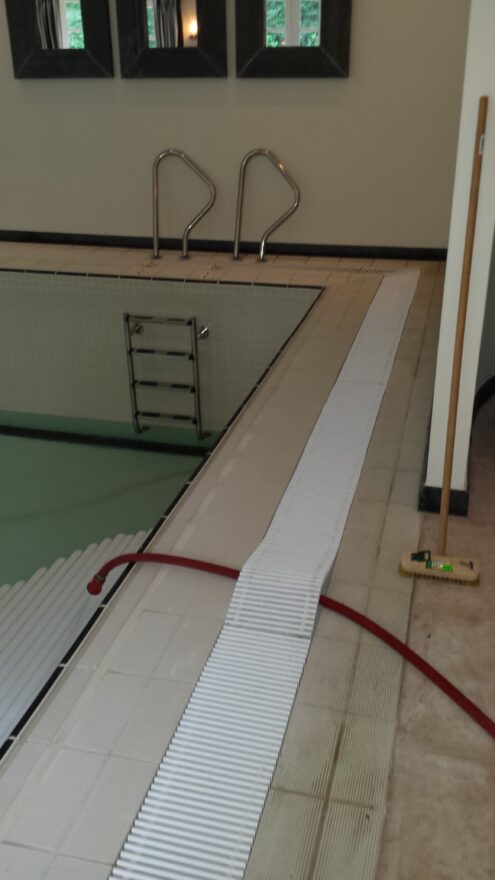 Zijkant van het binnenzwembad met trapleuning, ladder, wit rooster en een rode slang.