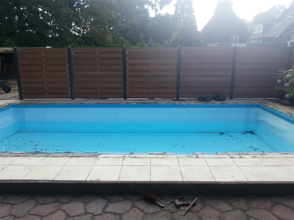 Zwembad met oude folie en lichte randtegels tegen een houten schutting, voorafgaand aan de renovatie.