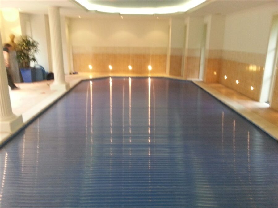 Binnenzwembad in Bussum met gesloten blauwe lamellenafdekking tussen witte zuilen en tegelwanden met wandverlichting.