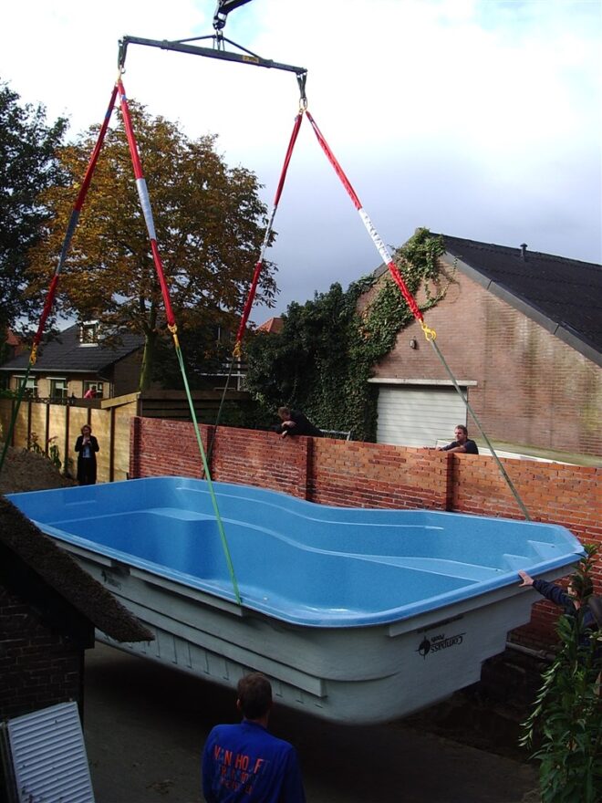 Het bassin wordt langs een woning gehesen aan kabels, vlak boven de grond.