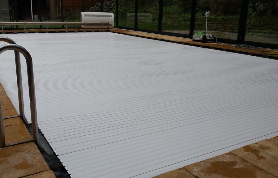 Gesloten witte lamellenafdekking over het binnenzwembad, met metalen trapleuning en glazen wand erachter.