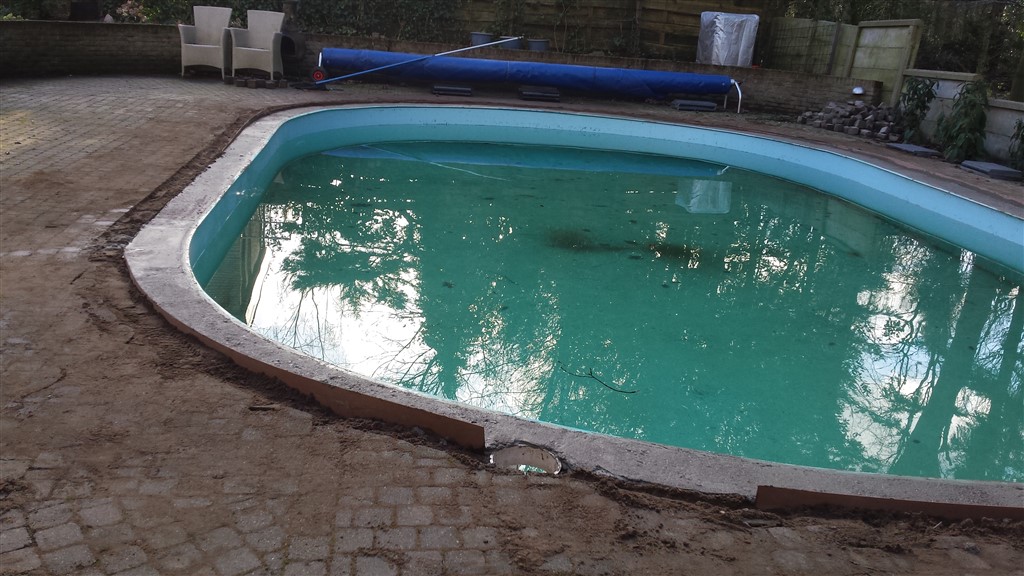 Zwembad met groenig water en een deels losgehaalde rand, met opgerolde blauwe afdekking op de achtergrond.