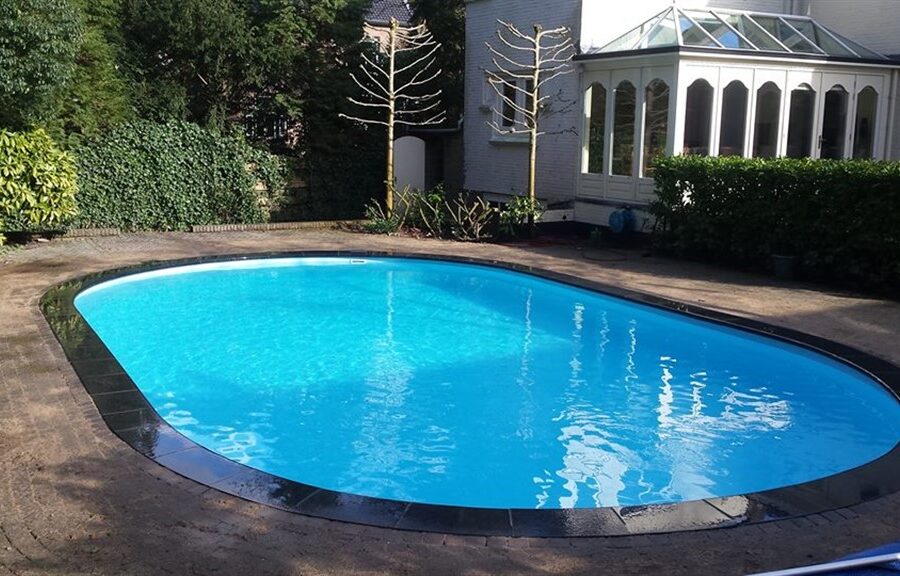 Ovaal zwembad in Baarn met helderblauw water en een strakke zwarte rand, tuin en serre erachter.