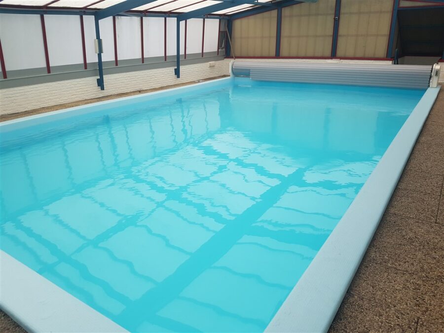 Binnenzwembad met lichtblauw water en lamellenafdekking aan de kopse kant, in een hal met lichtdoorlatend dak.