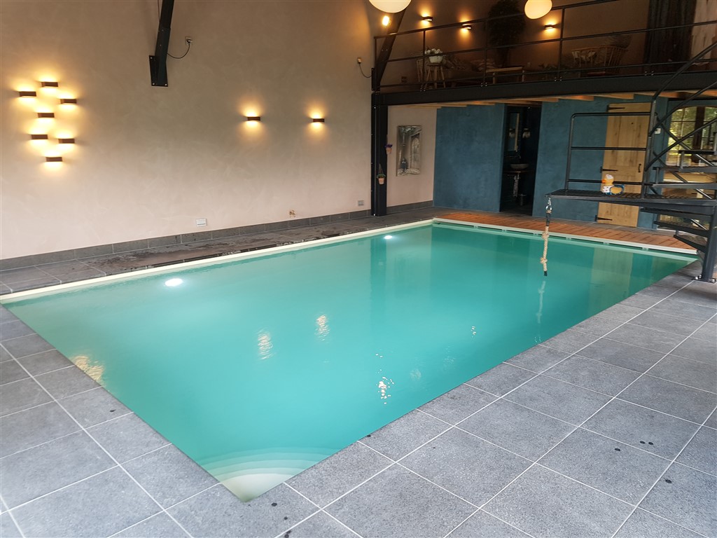 Afgewerkt binnenzwembad met turquoise water, grijze stenen vloer en wandverlichting in een ruimte met houten balken.