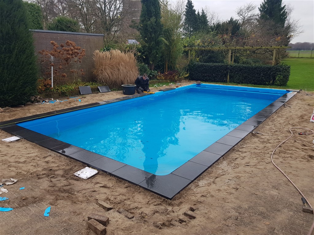 Gevuld zwembad met donkere randstenen op zand, nog voordat het terras is aangelegd.