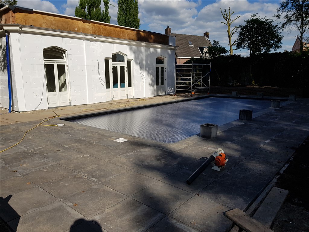 Terras bijna klaar rond het zwembad met gesloten afdekking, naast een wit gebouw.