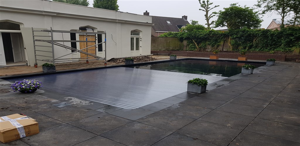 Zwembad met gesloten afdekking in een afgewerkt terras van donkere tegels, naast een wit gebouw.