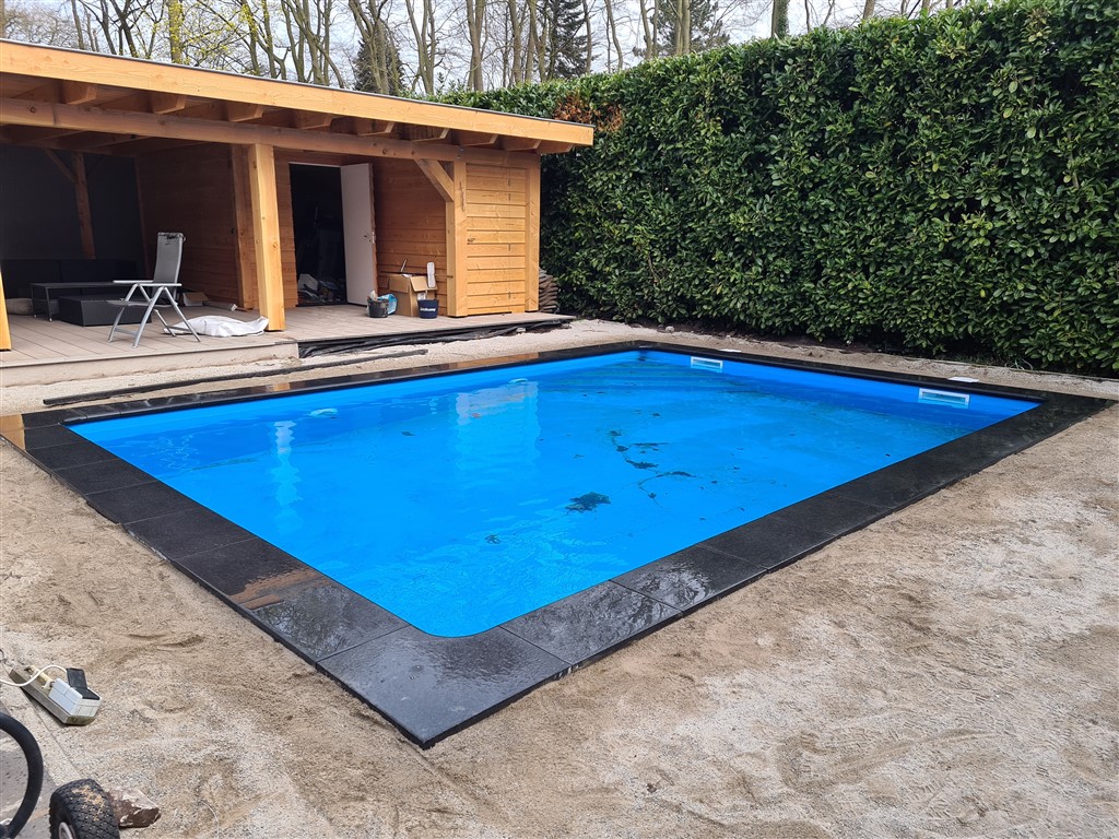 Het gevulde bassin met donkere natuurstenen rand, voor het houten bijgebouw en de haag.
