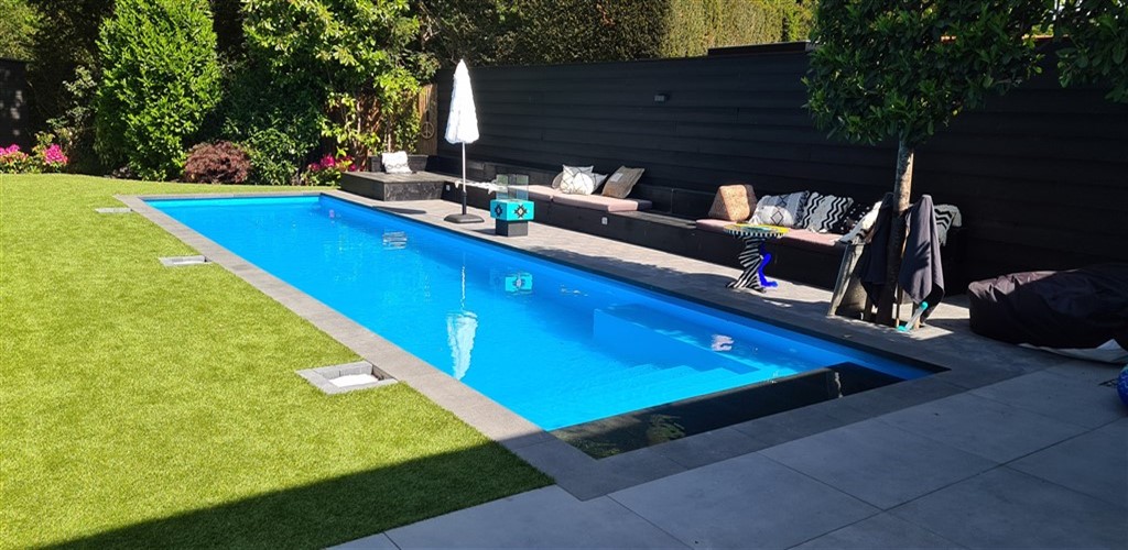 Open zwembad met blauw water langs het gazon, met een zitbank bij de zwarte schutting.