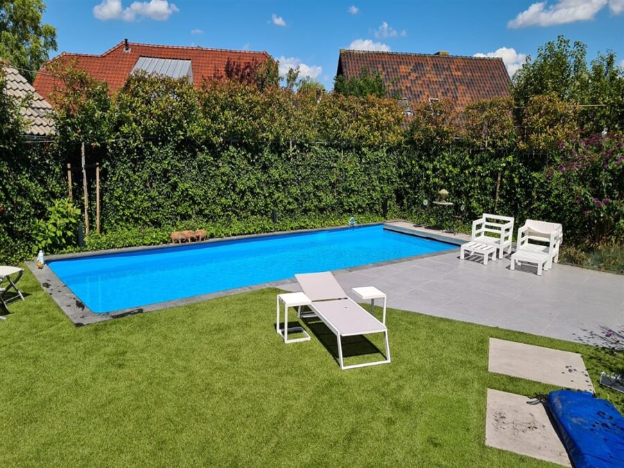 Helderblauw zwembad in een tuin met gazon, hagen en witte ligbedden in Benthuizen.