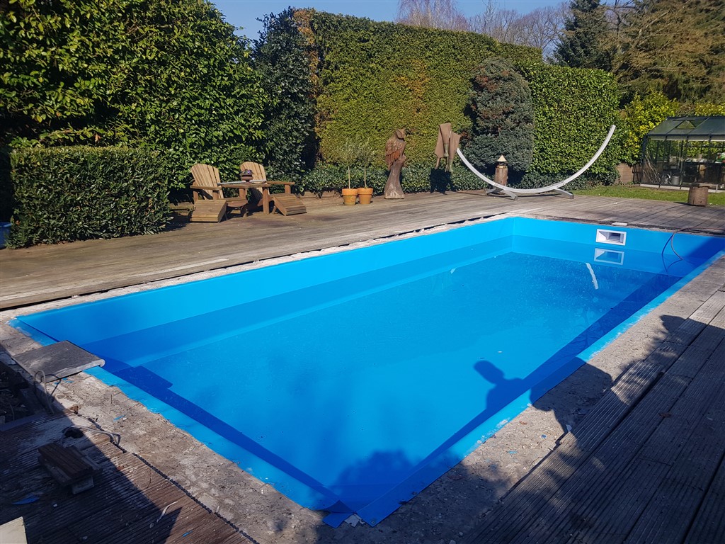 Het bassin met glanzende blauwe folie, met de hoge haag en tuinstoelen erachter.