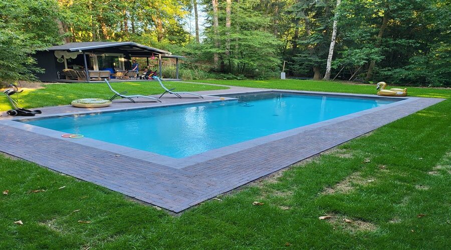 Opgeleverd L-vormig bassin met turquoise water in een gazon, met een houten tuinhuis erachter.