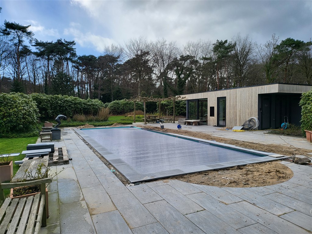 Het bassin onder een grijze afdekking, met terras van lichte tegels en een tuinhuis.