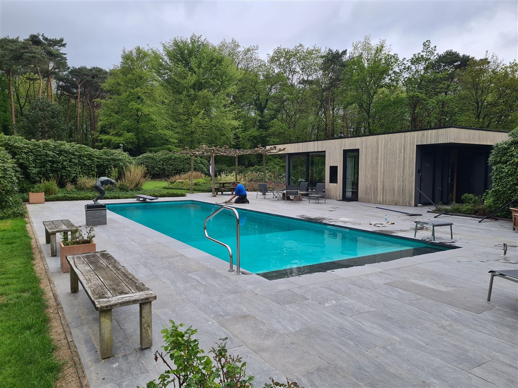 Het afgewerkte bassin met turquoise water, ladder en bankjes, met het houten tuinhuis op de achtergrond.